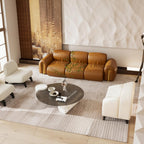 A modern living room scene featuring a tan leather sofa with green leaf embroidery on the seat cushion, styled with cream armchairs, a round dark coffee table, and a striped area rug.