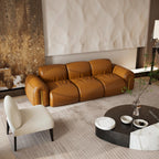 A modern three-seater leather sofa in a warm camel brown color, styled in a contemporary living room with a cream accent chair, a round black and white coffee table, and a textured beige wall behind it.
