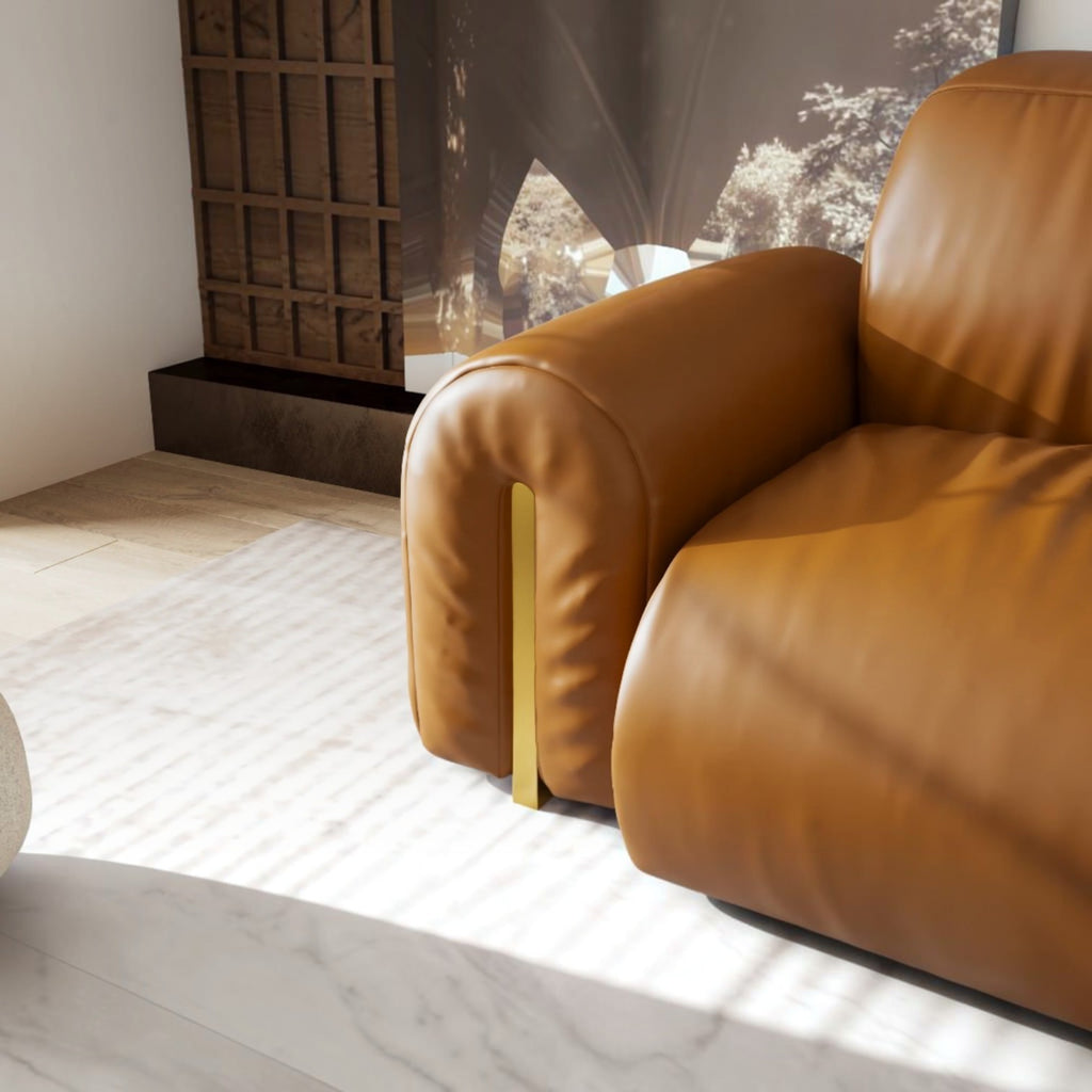 A close-up of a modern tan leather sofa with a distinctive vertical gold metal accent on the armrest, set in a contemporary living room with marble flooring and a textured wall.