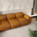 A modern three-seater sofa upholstered in warm camel-brown leather, shown in a minimalist living room setting with a textured light rug and a small potted plant.