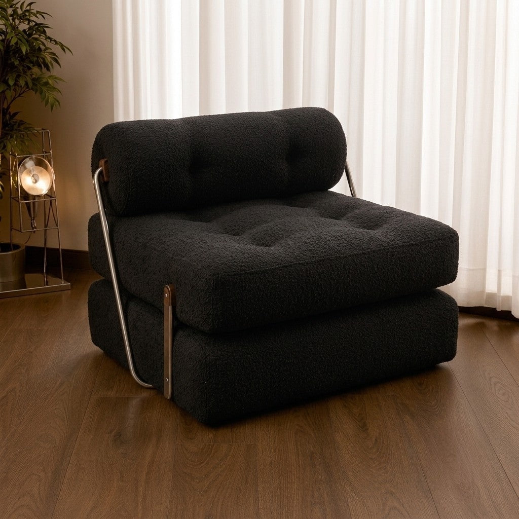 A black bouclé armchair with a tufted seat cushion and curved metal legs, shown in a styled room with wood flooring, white curtains, and a side table with a lamp.