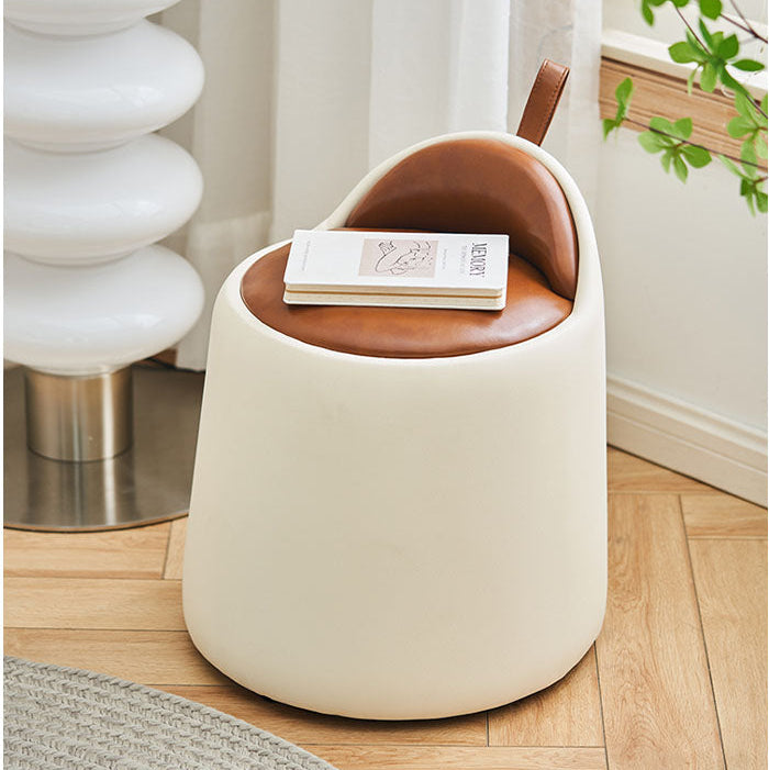 Small round stool with brown cushioned top and white base in a room with a plant and lamp.