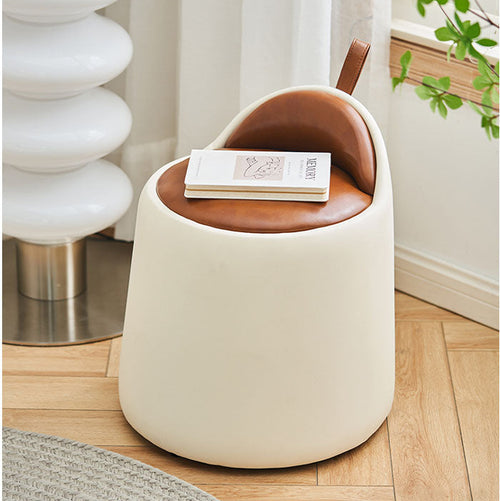 Small round stool with brown cushioned top and white base in a room with a plant and lamp.