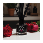 Diffuser with black reeds and a label, surrounded by red roses on a blurred background