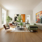 A bright, modern living room with light wood floors, featuring a sage green sofa, a brown leather lounge chair, black side tables, and two large abstract paintings on the walls.