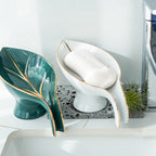 Teal leaf-shaped soap dish and white ceramic soap dish with soap on a bathroom counter.