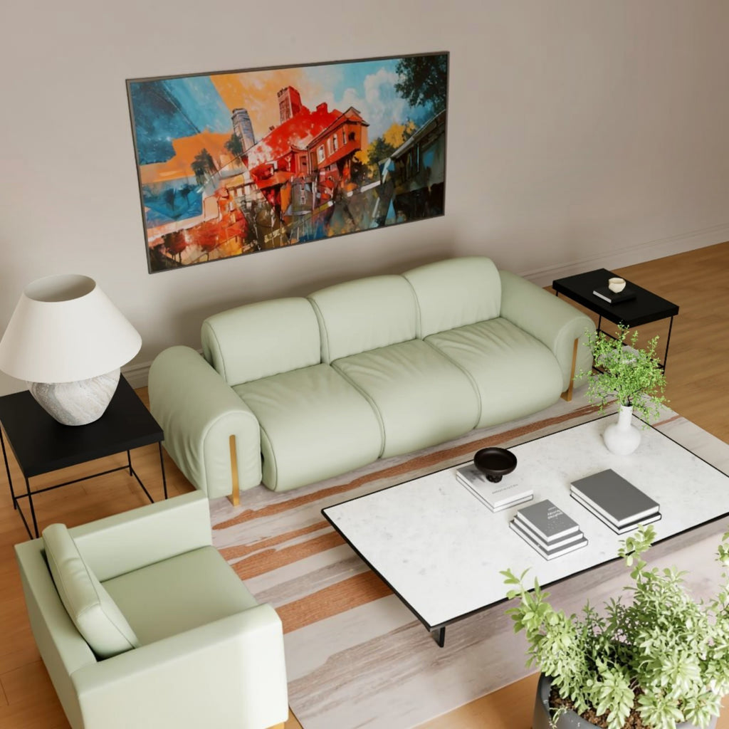 A modern living room setup featuring a light sage green three-seater sofa and a matching armchair, styled with a white marble coffee table, black side tables, a lamp, and a large colorful painting on the beige wall.