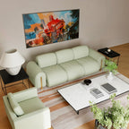 A modern living room setup featuring a light sage green three-seater sofa and a matching armchair, styled with a white marble coffee table, black side tables, a lamp, and a large colorful painting on the beige wall.