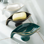 Two ceramic leaf-shaped soap dishes with a bar of soap on a tiled bathroom floor.