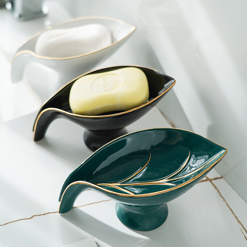Two ceramic leaf-shaped soap dishes with a bar of soap on a tiled bathroom floor.