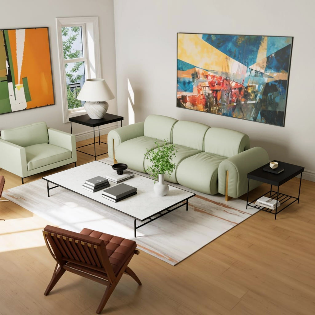 A modern living room featuring a light sage green modular sofa as the main product, styled with a white coffee table, black side tables, a brown leather chair, and colorful abstract wall art on a light wood floor.