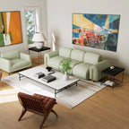 A modern living room featuring a light sage green modular sofa as the main product, styled with a white coffee table, black side tables, a brown leather chair, and colorful abstract wall art on a light wood floor.