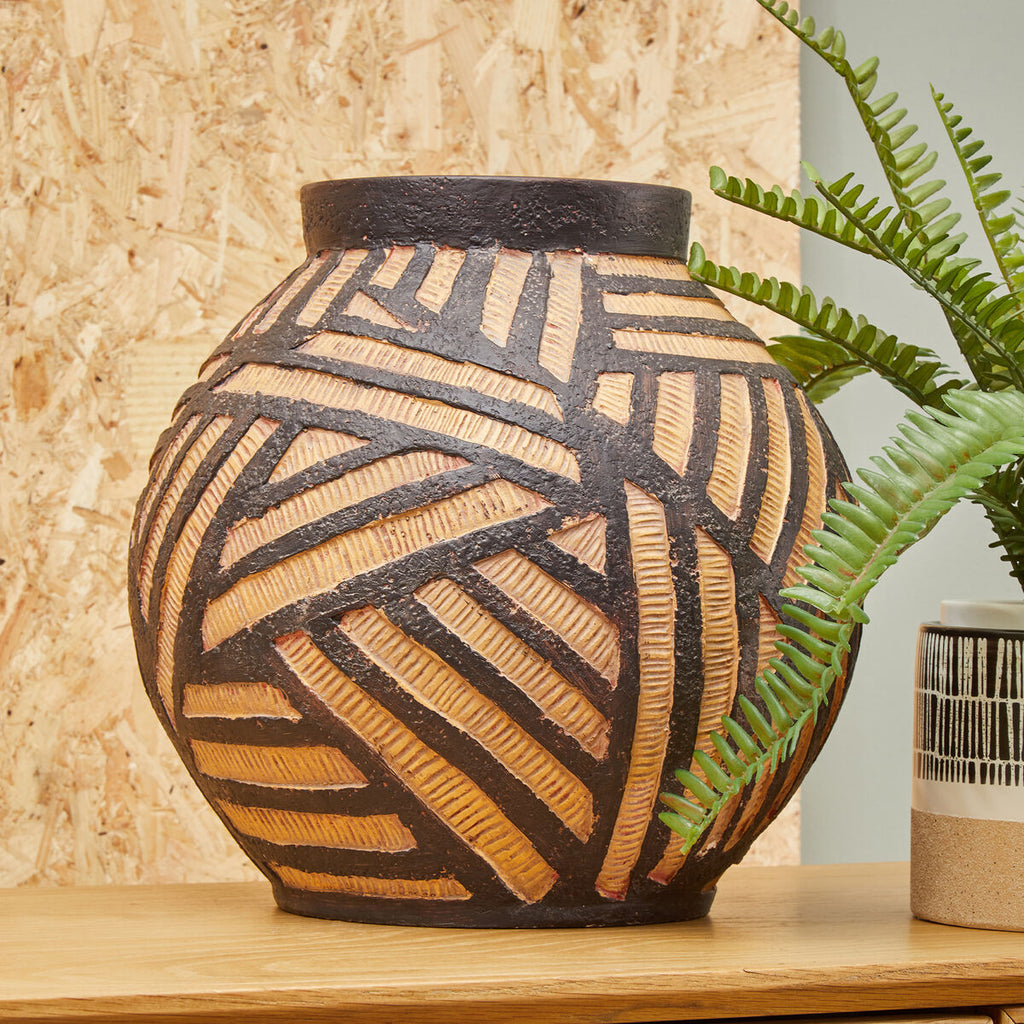 Decorative ceramic vase with geometric patterns on a wooden surface, with a plant in the background.