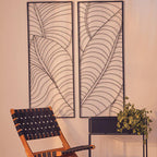 Two decorative wall panels with leaf patterns on a beige wall, with a chair and plant in the foreground.