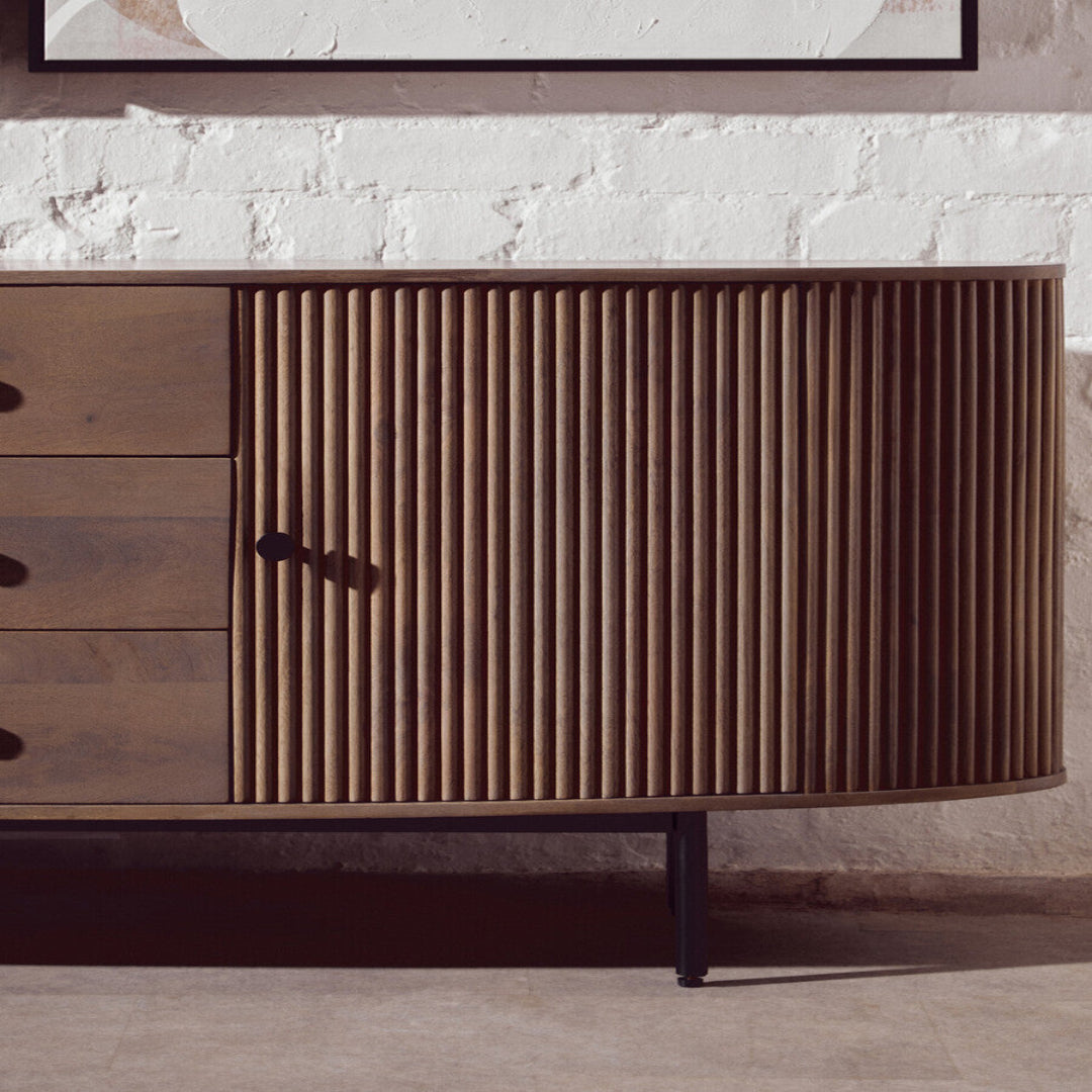 Wooden sideboard with a textured door and drawers against a light stone wall.