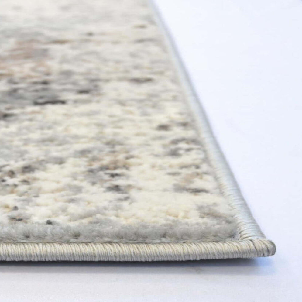 Close-up of a textured area rug corner showing a cream and gray marbled pattern with a light gray twisted border on a white surface.