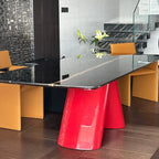 A modern dining table with a glossy black glass top and a bold, sculptural bright red base, styled in a contemporary dining room with orange chairs and a glass staircase railing.