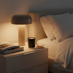 Nighstand with lamp, book, and candle next to a bed in a dimly lit room