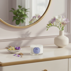 Candle with a decorative label on a table next to a vase with flowers and a round mirror.