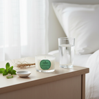 Candle with a green label on a bedside table next to a glass of water and a small dish with herbs.