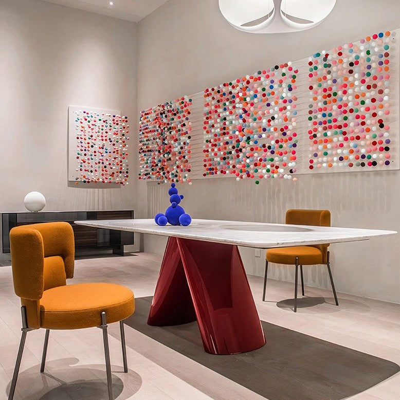 A modern dining room scene featuring a white rectangular table with a glossy red sculptural pedestal base, surrounded by two mustard yellow upholstered chairs. The room has light beige walls decorated with three large, colorful circular dot art installations and a matching smaller piece on the left.