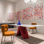 A modern dining room scene featuring a white rectangular table with a glossy red sculptural pedestal base, surrounded by two mustard yellow upholstered chairs. The room has light beige walls decorated with three large, colorful circular dot art installations and a matching smaller piece on the left.