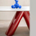A modern table with a glossy red V-shaped base and a white marble top, styled with a blue sculptural bear figurine on a light wood floor.