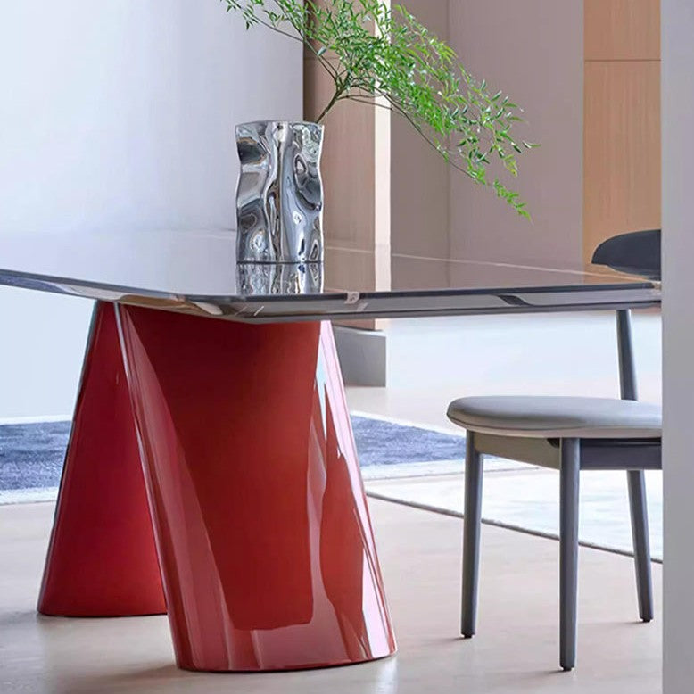 A modern dining table with a glossy red sculptural base and a clear glass top, styled in a minimalist dining room with a chair and a silver vase.