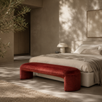 A modern bedroom scene with a bed dressed in neutral beige bedding, a deep red velvet bench at the foot, textured plaster walls, and dappled sunlight casting leaf shadows across the room.