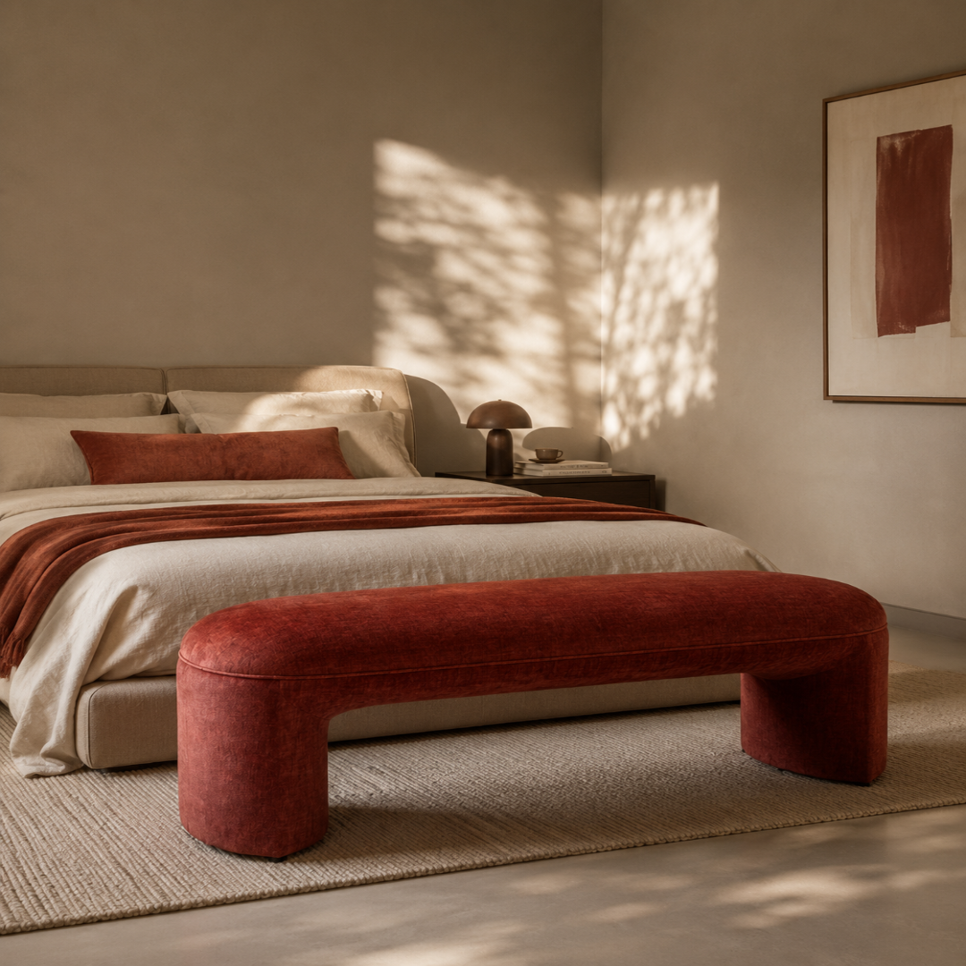 A modern bedroom scene featuring a low, wide upholstered bench in a rust-red velvet fabric at the foot of a bed. The bed is dressed in neutral beige and cream linens with a matching rust-red throw blanket.