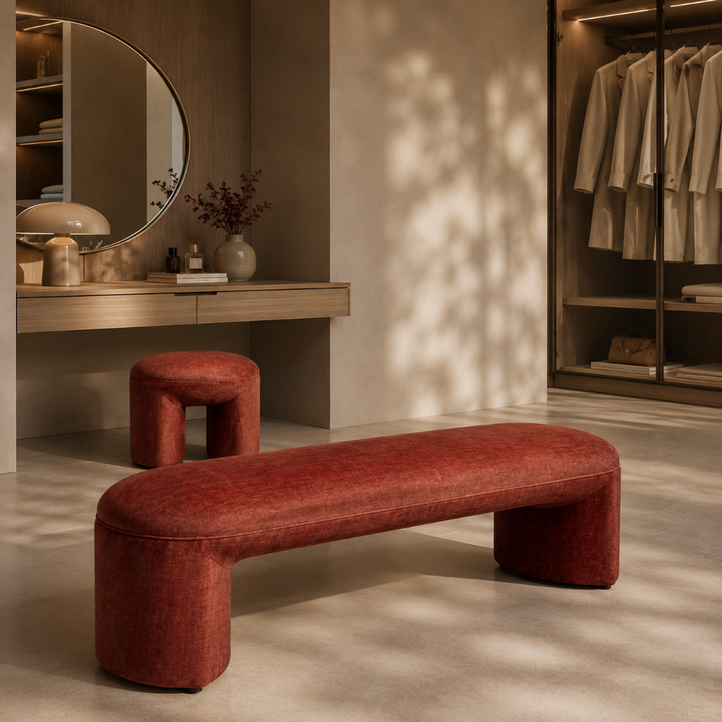 A modern curved upholstered bench in textured rust red fabric, positioned in a warm, stylish dressing room or closet area with a matching small stool, vanity, mirror, and hanging garments in the background.