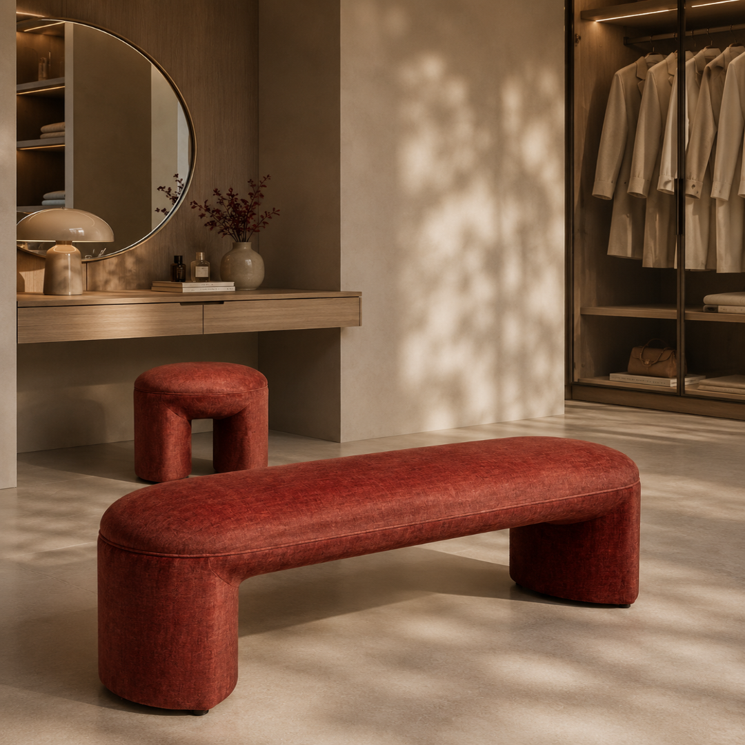 A modern curved upholstered bench in textured rust red fabric, positioned in a warm, stylish dressing room or closet area with a matching small stool, vanity, mirror, and hanging garments in the background.