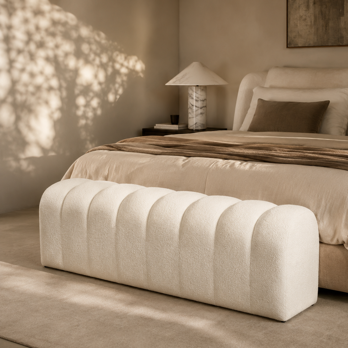 A modern bedroom scene featuring a tufted, barrel-shaped upholstered bench in a warm off-white fabric at the foot of a bed. The bed is dressed in neutral beige and brown linens, with a matching lamp on a side table and dappled sunlight casting shadows on the wall.