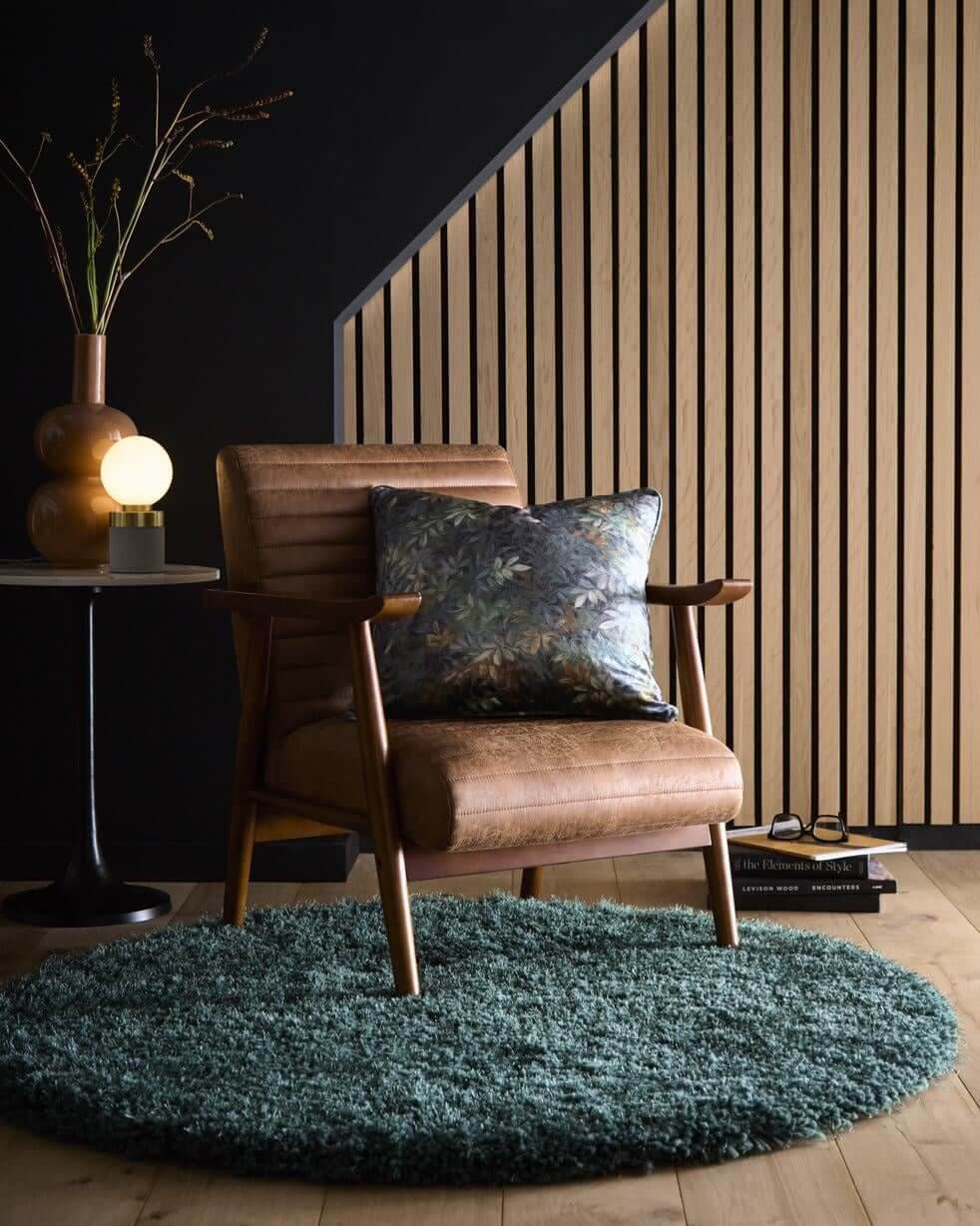 Wooden armchair with a decorative pillow on a green rug in a room with a dark wall and wooden slat panel.