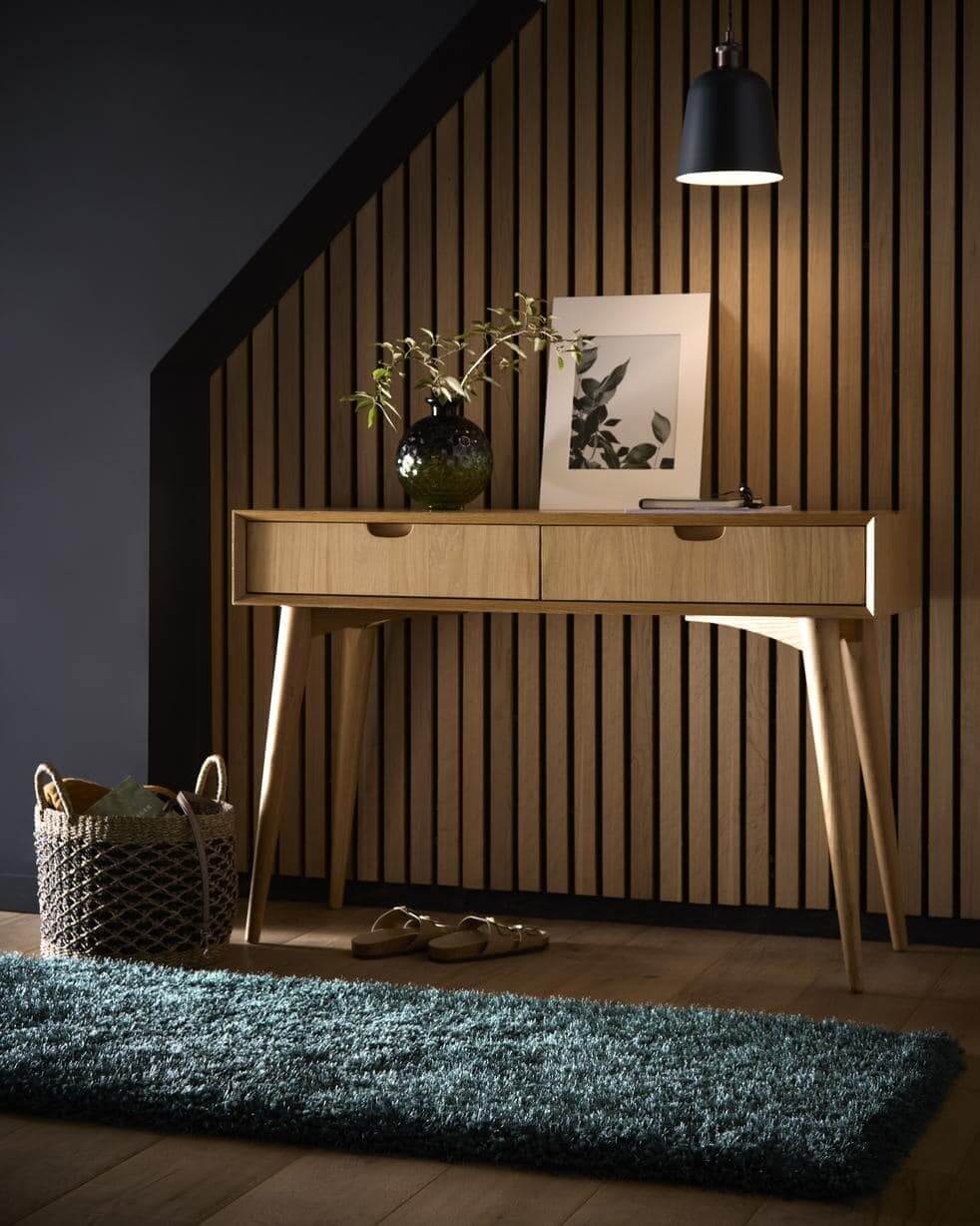 Wooden console table against a slatted wall with a plant and framed picture on top, in a room with a blue rug and shoes.