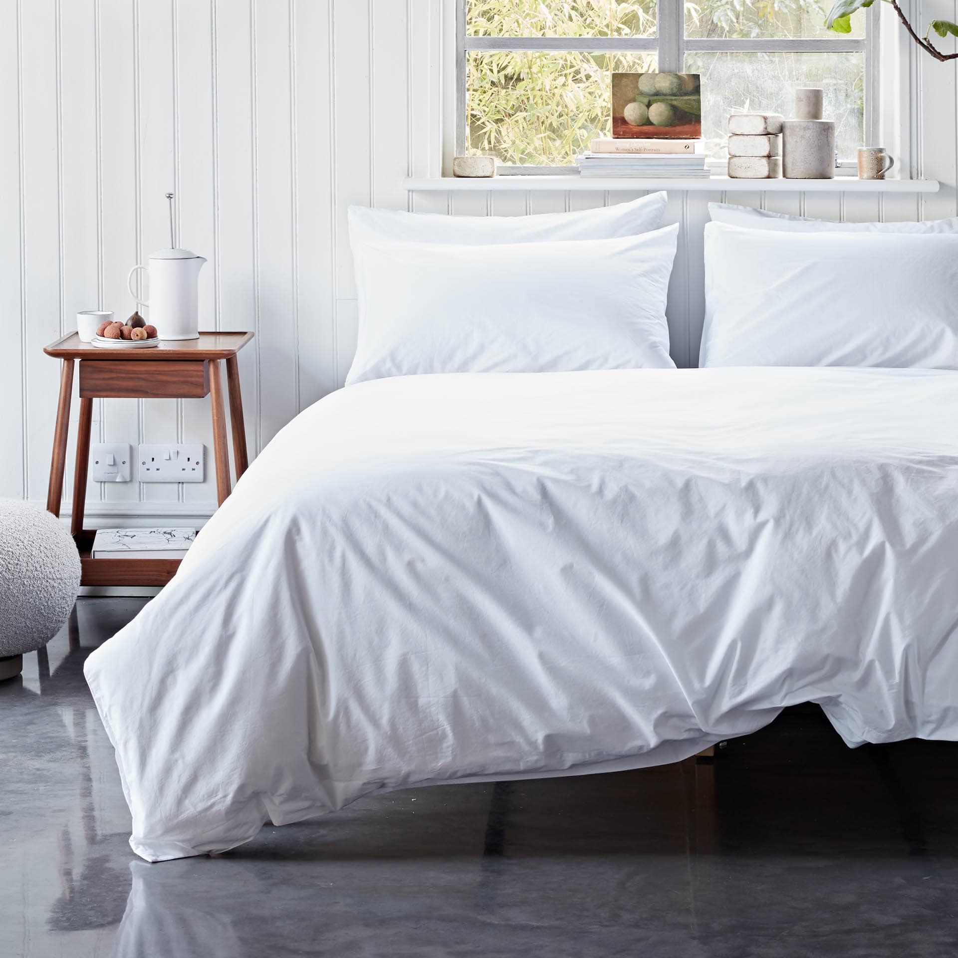 White bedding set on a bed in a bright bedroom with a wooden side table.