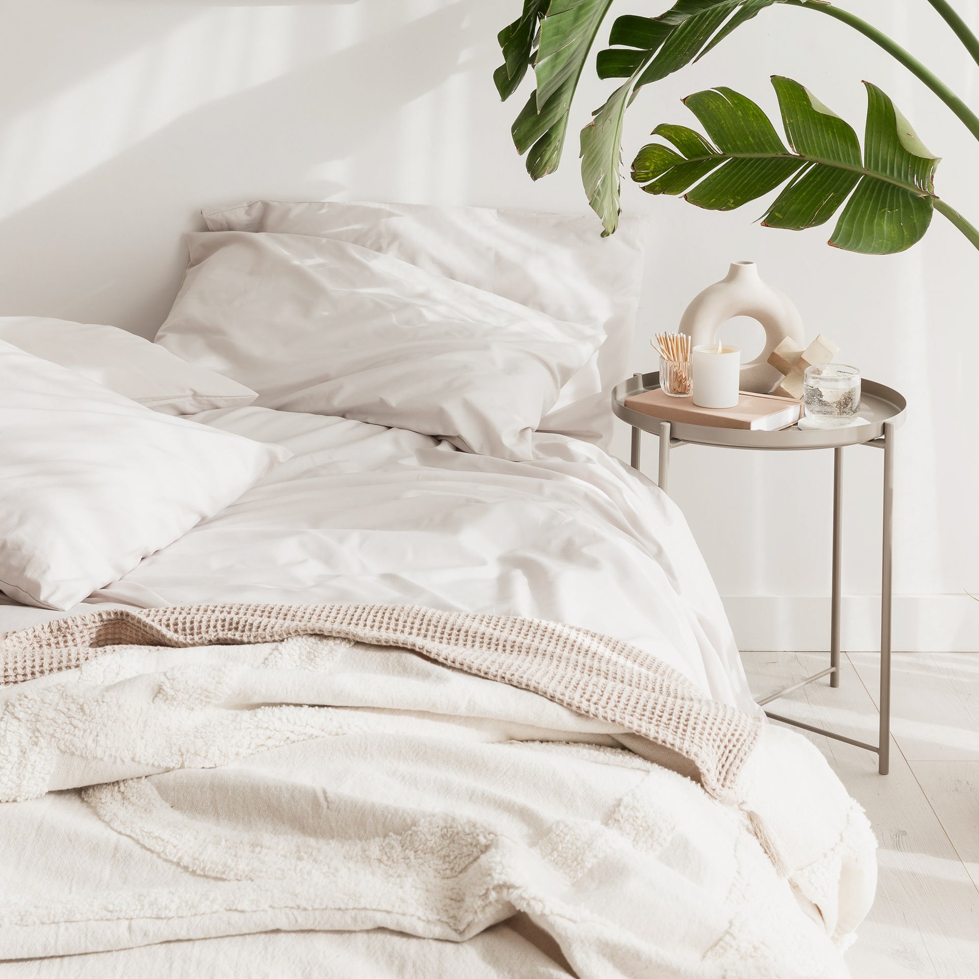 Neatly made bed with white bedding and a side table with a plant and candles in a bright room.