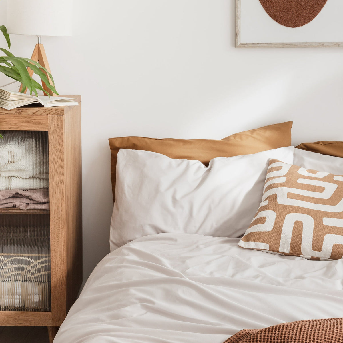 Bedroom with a bed featuring white bedding and brown pillows, next to a wooden nightstand with books and a plant.