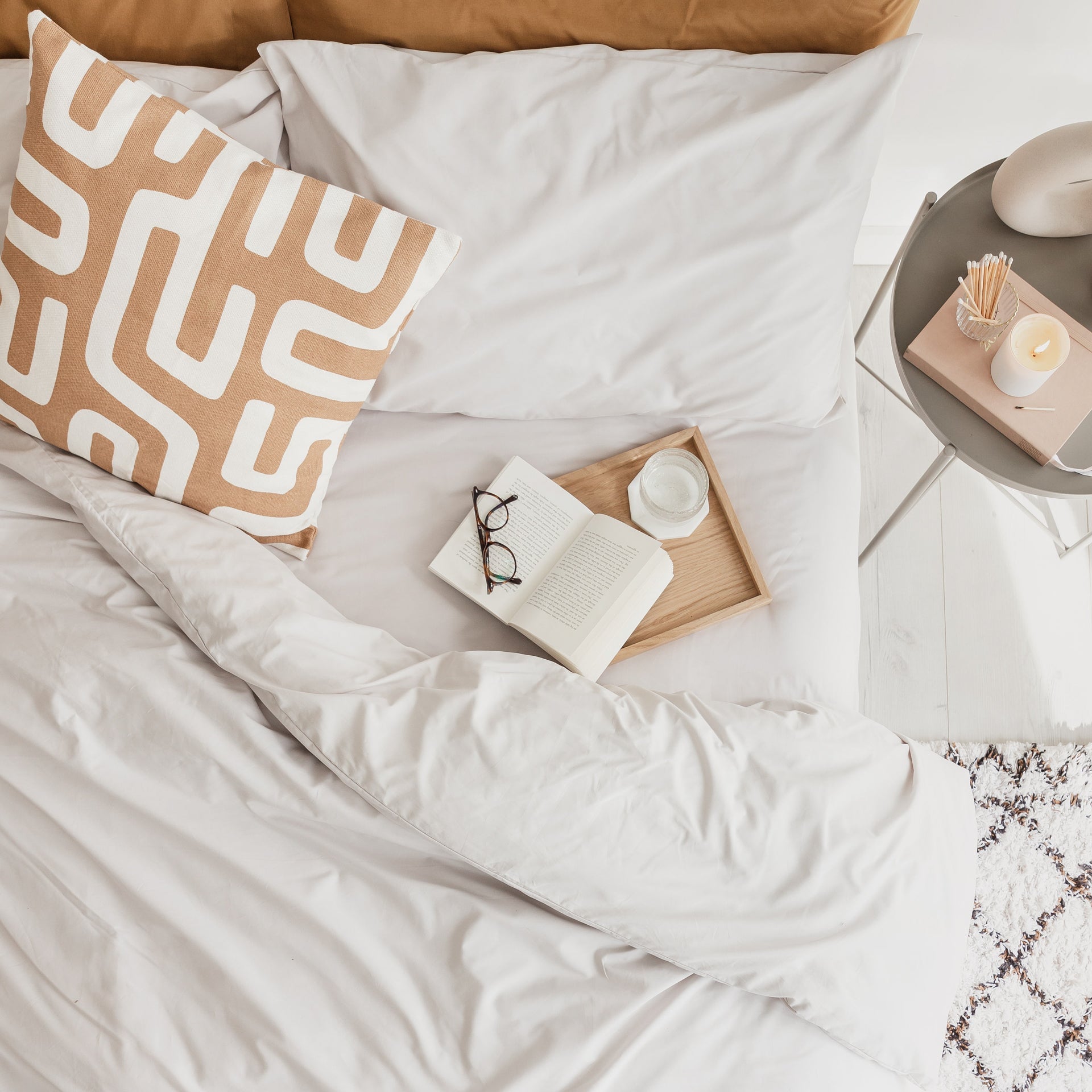 Neatly made bed with a decorative pillow and small wooden tray on a light-colored surface.