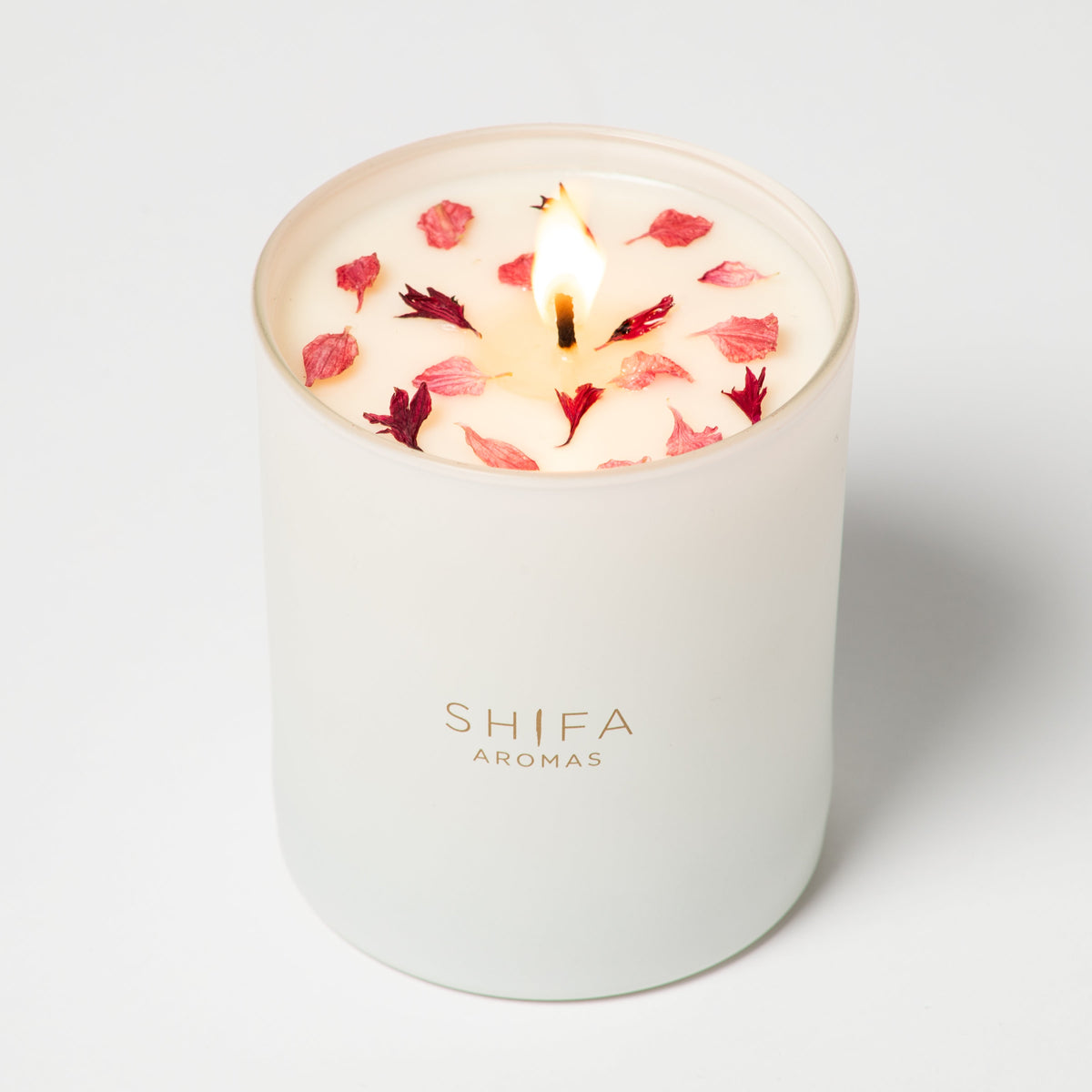 Candle with floral petals in a white container labeled 'SHIFA AROMAS' on a light gray background