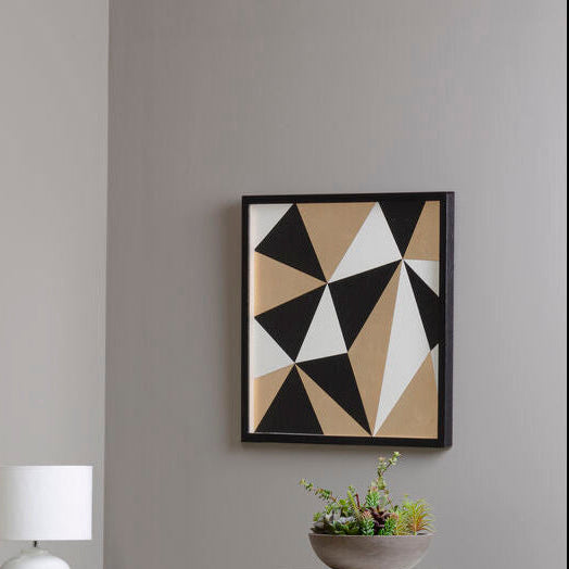 Geometric abstract art piece on a gray wall with a lamp and plant in the foreground.