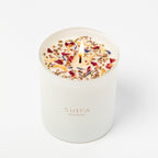 White candle with colorful dried flowers on a white background, branded 'Shifa Aromas'.