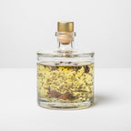 Clear glass bottle with gold cap containing a yellow liquid and floating herbs on a white background