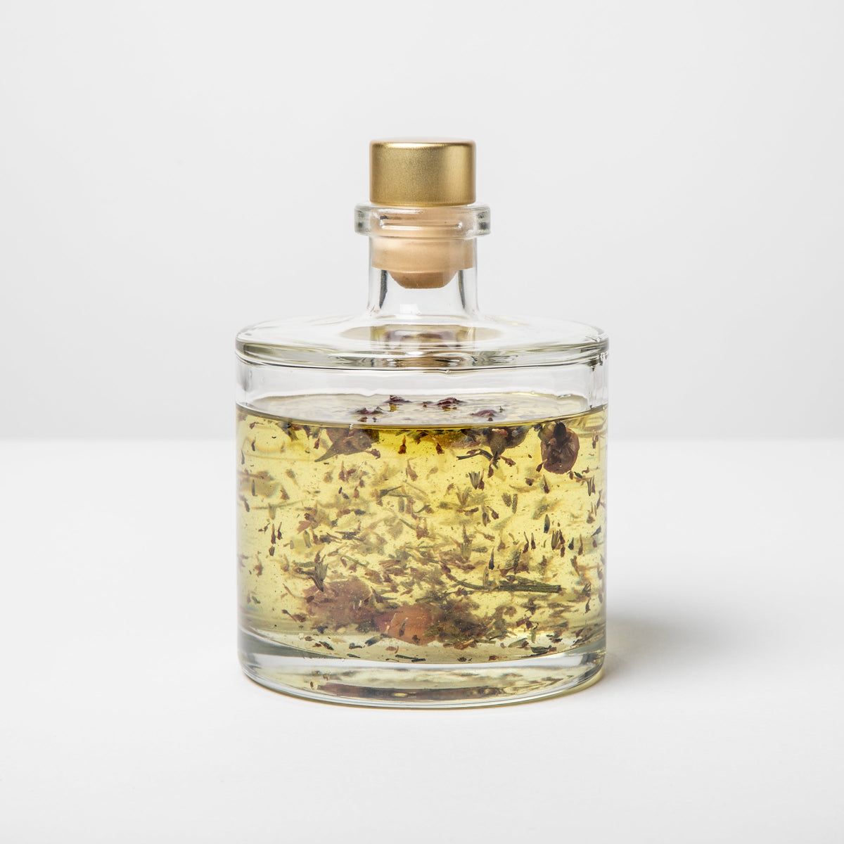 Clear glass bottle with gold cap containing a yellow liquid and floating herbs on a white background