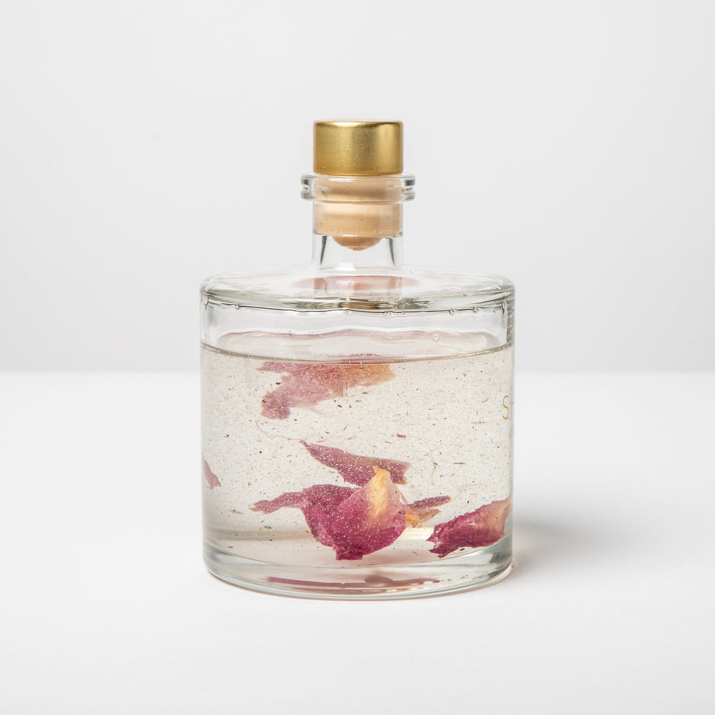 Clear glass bottle with gold cap containing water and pink petals on a white background