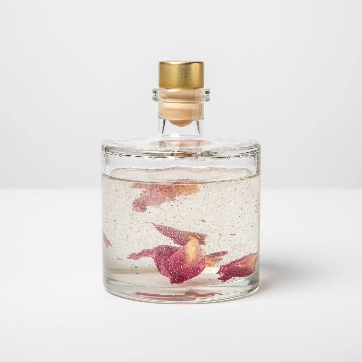 Clear glass bottle with gold cap containing water and pink petals on a white background