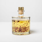 Glass bottle with gold cap containing a yellow liquid with red and brown bits on a white background
