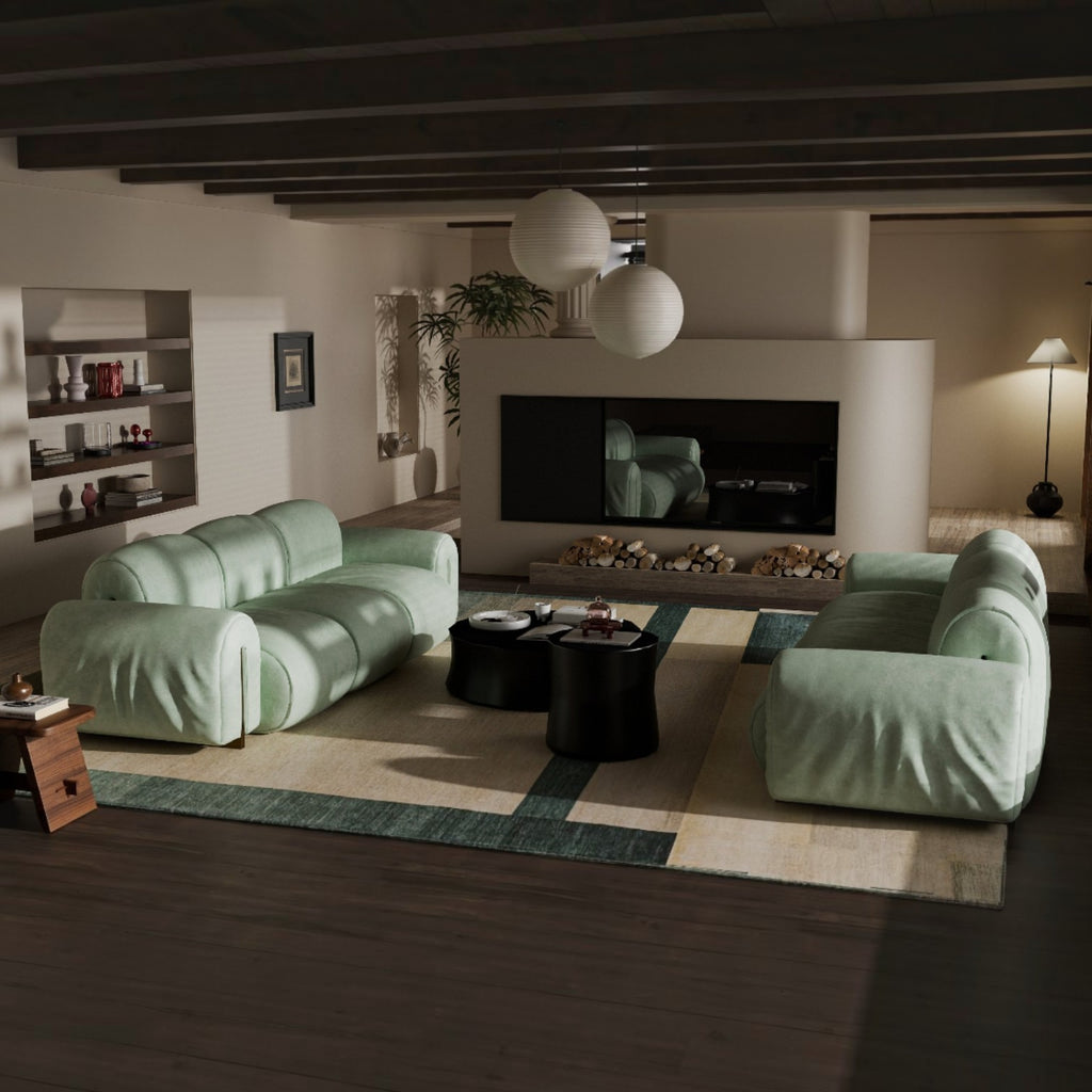 A modern living room featuring two oversized, curved light green sofas arranged on a geometric area rug. The room has dark wood ceiling beams, a built-in fireplace with a black surround, and two white spherical pendant lights hanging above.