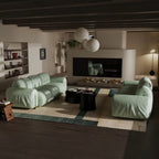 A modern living room featuring two oversized, curved light green sofas arranged on a geometric area rug. The room has dark wood ceiling beams, a built-in fireplace with a black surround, and two white spherical pendant lights hanging above.