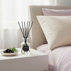 Diffuser with black sticks on a nightstand next to a bed with white pillows.
